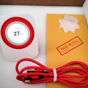 Griftmeter Baseline shell in its shipping box with the dial lit and showing a live reading, beside the envelope, stand and red cable