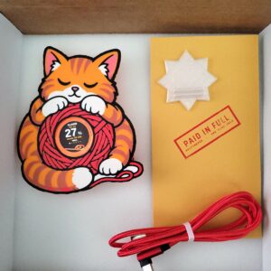 Griftmeter Cat shell in its shipping box with the dial lit and showing a live reading, beside the envelope, stand and red cable