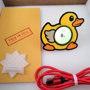 Griftmeter Rubber Duck shell in its shipping box with the dial lit and reading usage, beside the envelope, stand and red cable