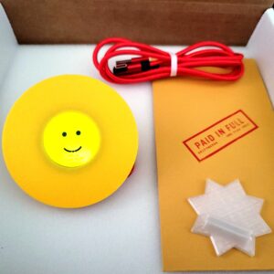 Griftmeter Emoji shell in its shipping box with the dial lit and showing a live reading, beside the envelope, stand and red cable