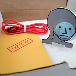 Griftmeter NPC shell in its shipping box with the dial lit and showing a live reading, beside the envelope, stand and red cable