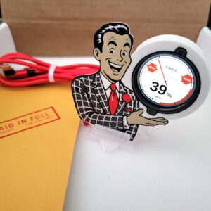 Griftmeter Salesman shell in its shipping box with the dial lit and showing a live reading, beside the envelope, stand and red cable