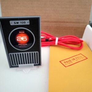 Griftmeter Sentinel shell in its shipping box with the dial lit and showing a live reading, beside the envelope, stand and red cable