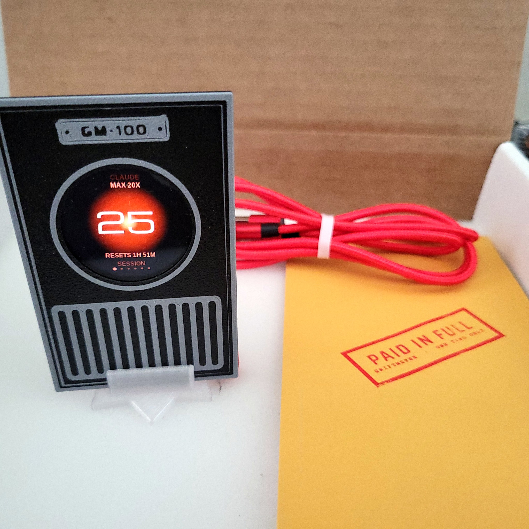 Griftmeter Sentinel shell in its shipping box with the dial lit and showing a live reading, beside the envelope, stand and red cable