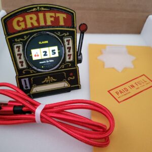 Griftmeter Slot Machine shell in its shipping box with the dial lit and showing a live reading, beside the envelope, stand and red cable
