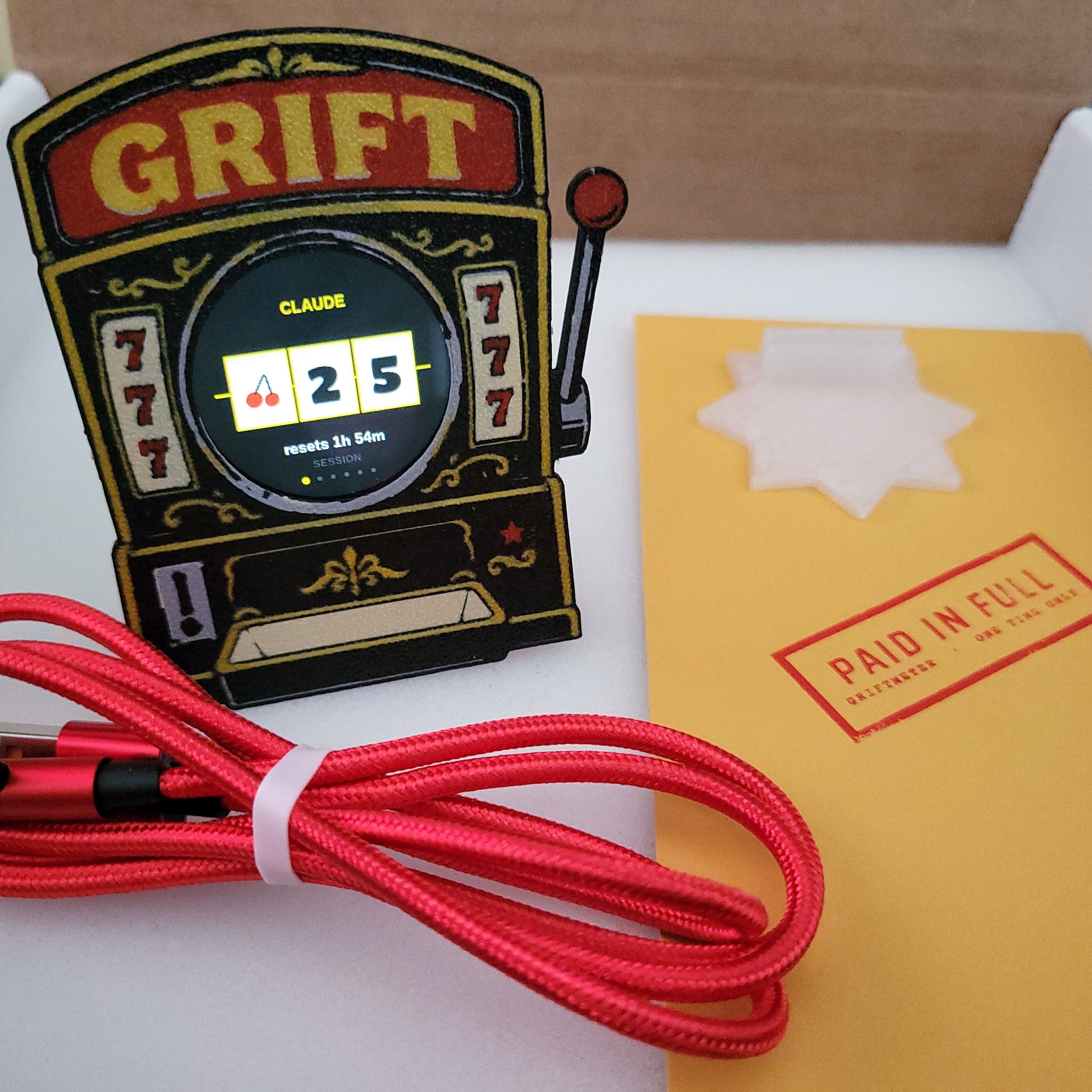 Griftmeter Slot Machine shell in its shipping box with the dial lit and showing a live reading, beside the envelope, stand and red cable