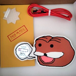 Griftmeter Spurdo shell in its shipping box with the dial lit and showing a live reading, beside the envelope, stand and red cable