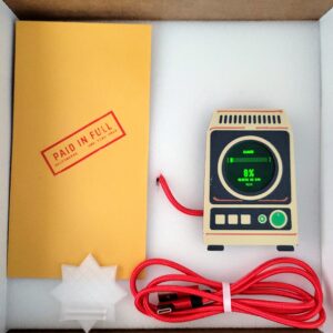 Griftmeter Terminal shell standing in its shipping box with the green screen lit and reading Claude usage, beside the envelope and red cable