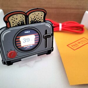 Griftmeter Toaster shell in its shipping box with the dial lit and showing a live reading, beside the envelope, stand and red cable
