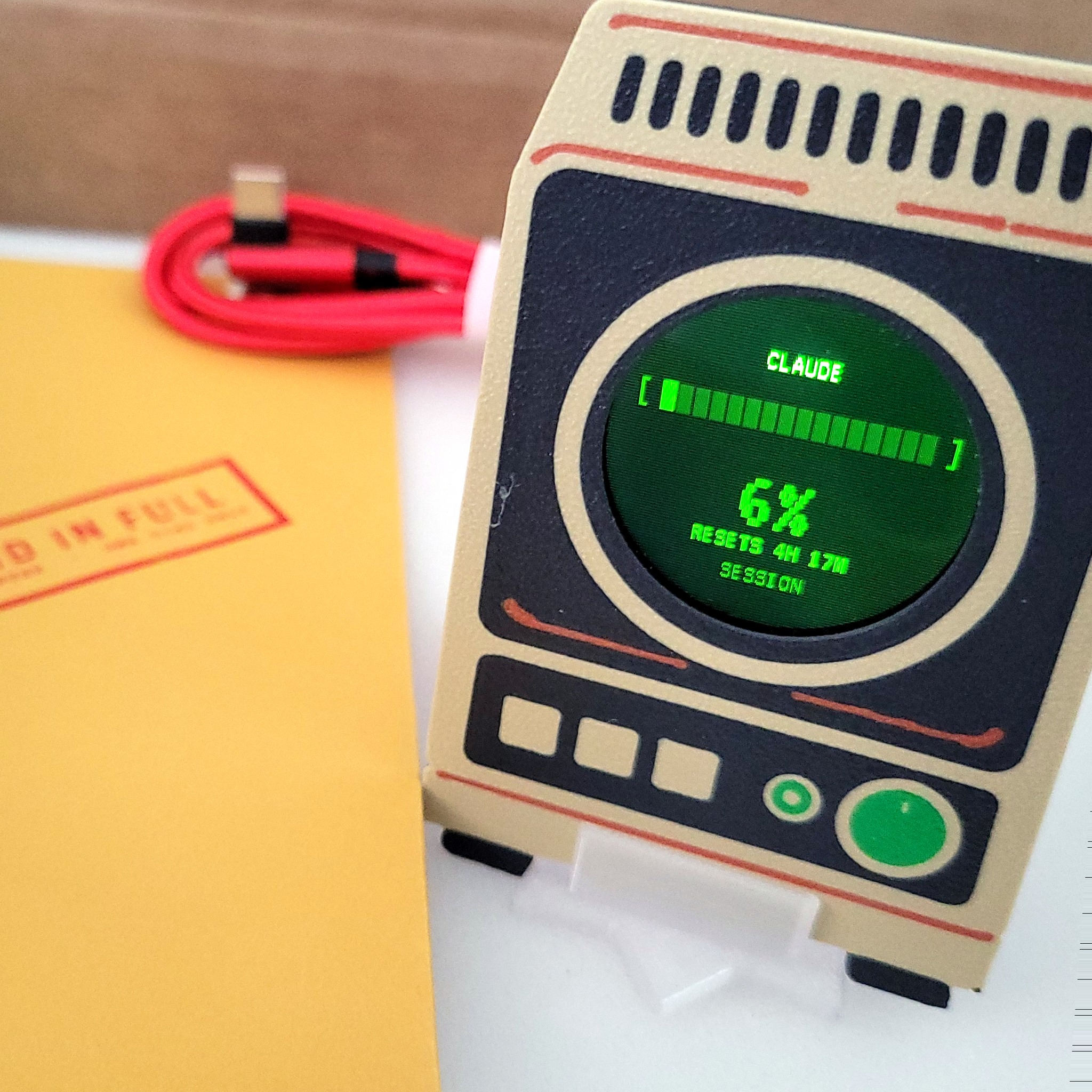 Griftmeter Starter Bundle: the Terminal shell on its stand in the box, screen lit and reading Claude usage, beside the envelope and red cable